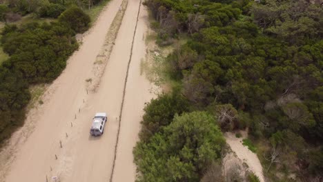 Luftaufnahme-Eines-Weißen-4x4-Radfahrzeugs,-Das-Bei-Sonnenlicht-Auf-Einem-Sandstrandweg-Zwischen-Wäldern-Fährt