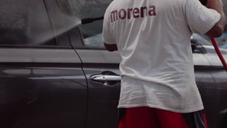 man in white shirt cleaning car with hose outdoors, daytime, urban setting, casual atmosphere