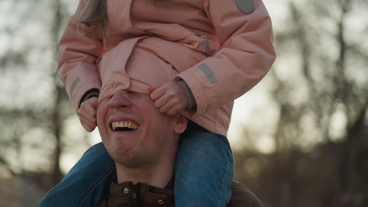 A joyful moment as a little girl in a pink cap and jacket sits on her dad's neck, playfully tucking his head inside her jacket. Both are laughing, capturing a heartwarming and playful interaction