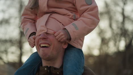 a joyful moment as a little girl in a pink cap and jacket sits on her dad's neck, playfully tucking his head inside her jacket. both are laughing, capturing a heartwarming and playful interaction