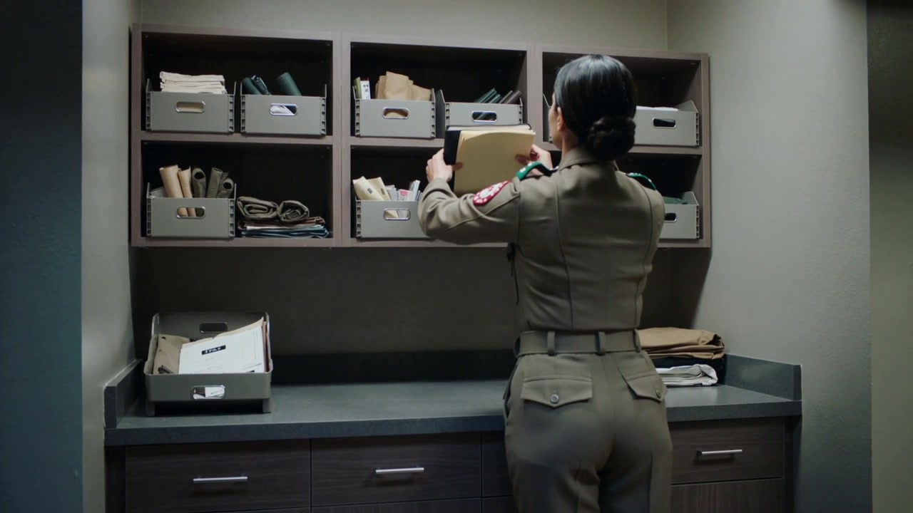 Picking up binder on counter, uniformed woman sorting manila files into labeled cubbies to organize