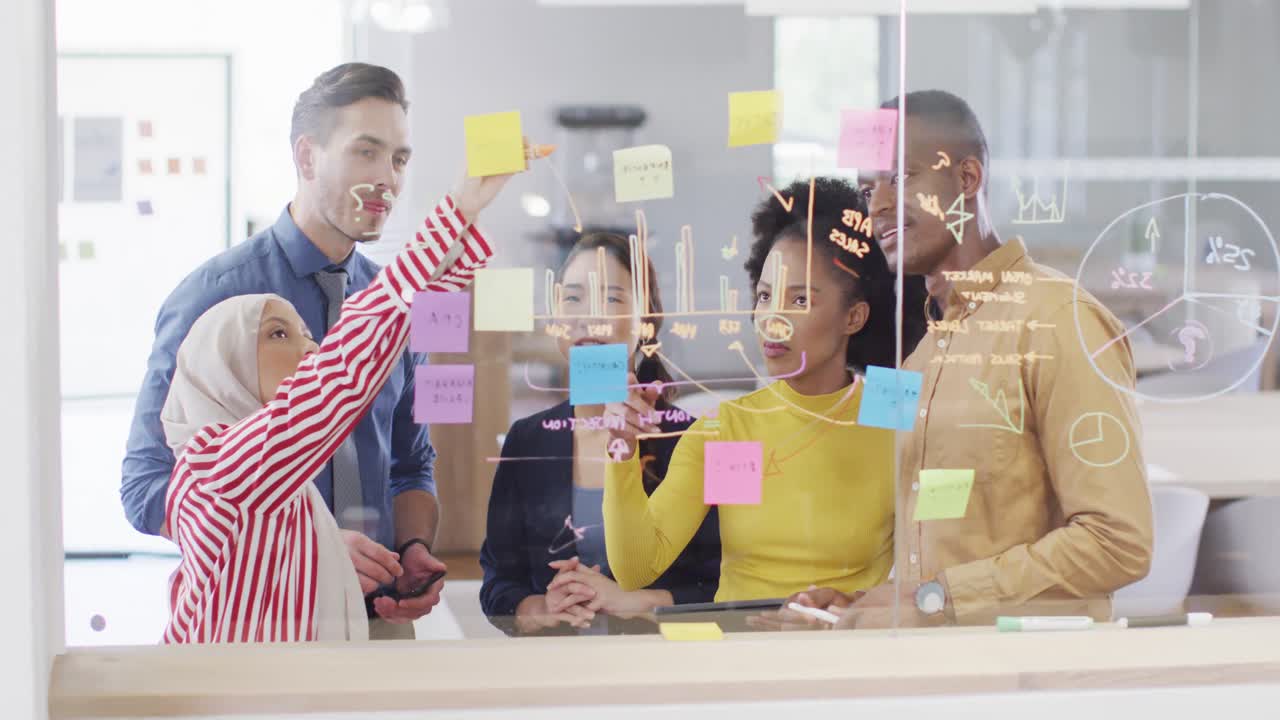 Group of diverse business people taking notes on glass wall and talking in office, slow motion