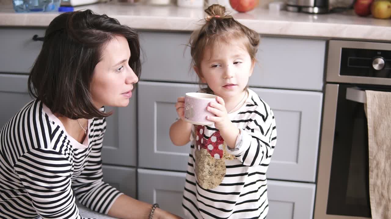 la madre le da a su hija una taza de agua, la niña bebe, la madre está sonriendo en la cocina