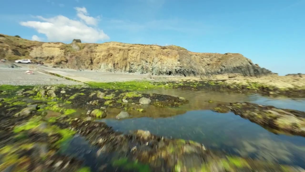 FPV-drone suser langs et taggete tangdekket rev med klipper og en parkeringsplass ved båtopptrekk synlig over det lyseblå havet