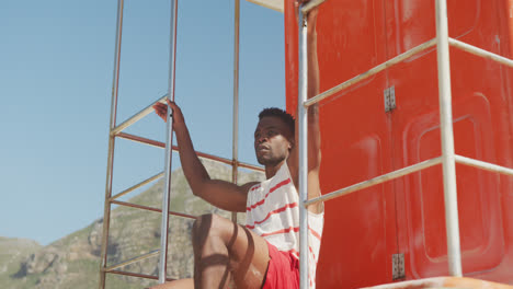 African-american-male-lifeguard-inspecting-on-tower-on-sunny-beach