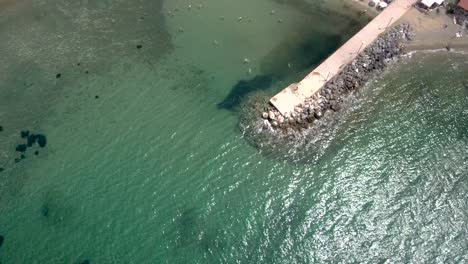 Vista-Aérea-De-Una-Pequeña-Playa-Turquesa-Con-Un-Muelle-En-El-área-De-Agia-Paraskevi-Halkidiki,-Grecia,-Movimiento-Hacia-Arriba-Y-Hacia-Adelante-Por-Drones