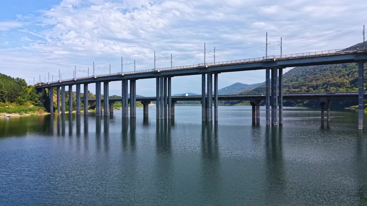 Ponte che attraversa un fiume con binari ferroviari sopra e riflessi nell'acqua sotto un cielo nuvoloso