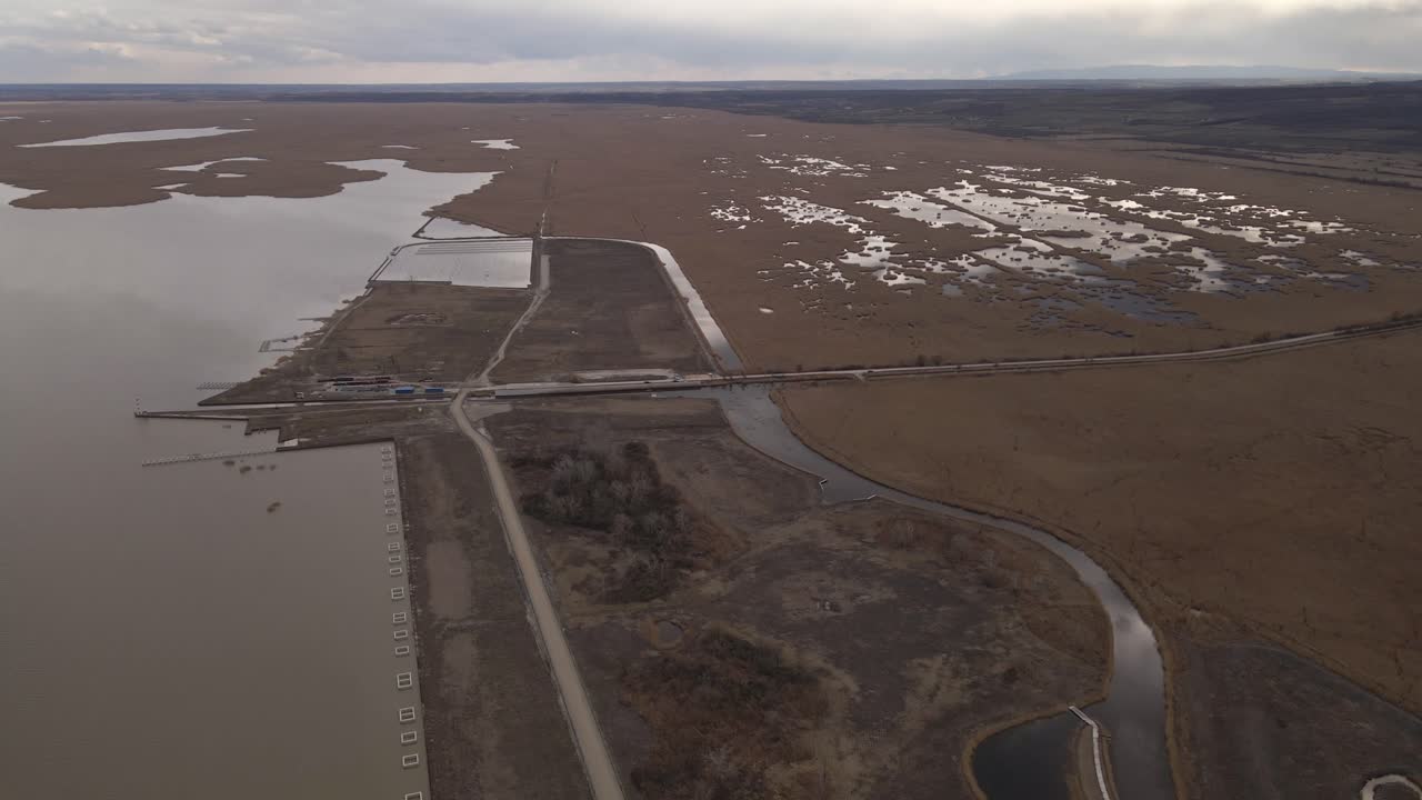 An aerial video of a lake and wetland area with a small harbor, roads, and infrastructure. The landscape features water channels, marshy patches, and a cloudy sky.