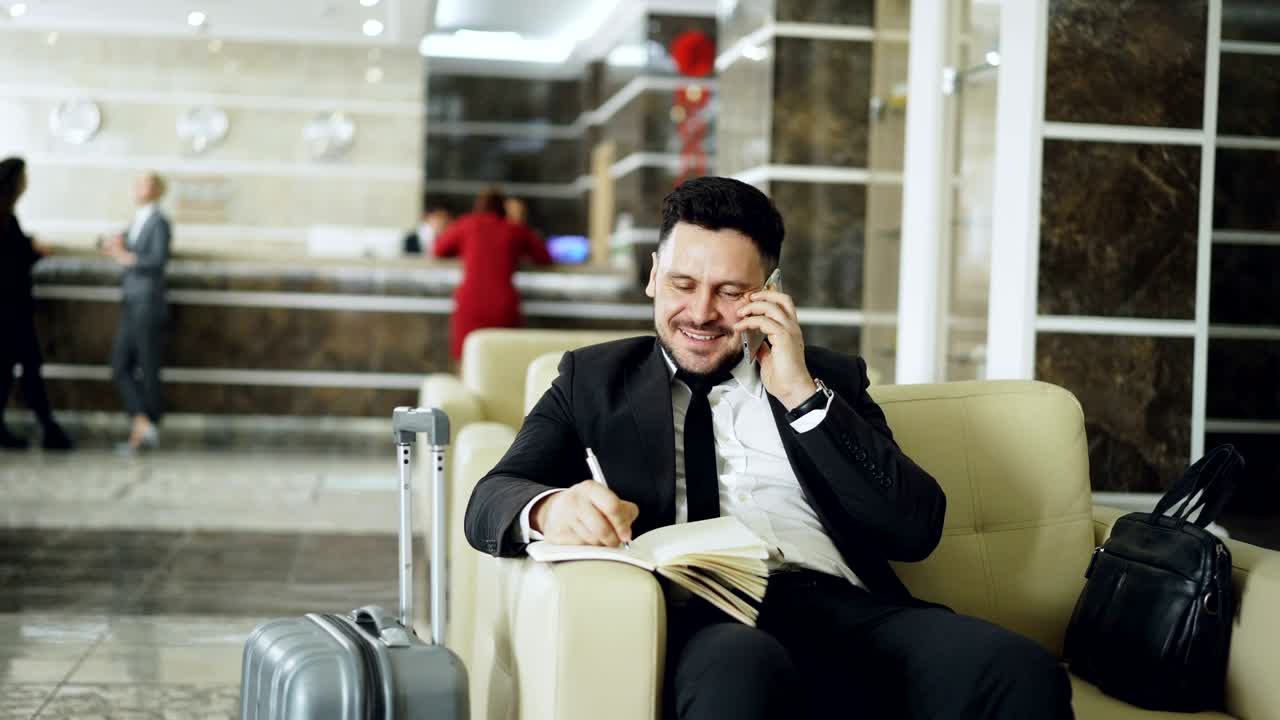 tomada de un hombre de negocios confiado y alegre hablando por teléfono móvil sonriendo y escribiendo notas en el bloc de notas mientras está sentado en el sillón en la recepción del hotel. concepto de viaje, negocios y personas