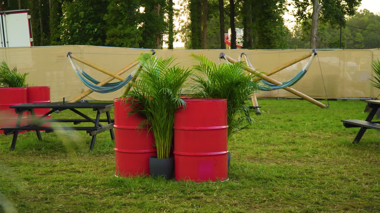 VIP area at the festival outdoor hidden from public area with hammocks red barrels surrounded by tropical plants many bench seats and tables on grassy field tent tipi in the background during sunset