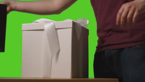 close up of man putting gift wrapped present in box into gift bag on table shot against green screen