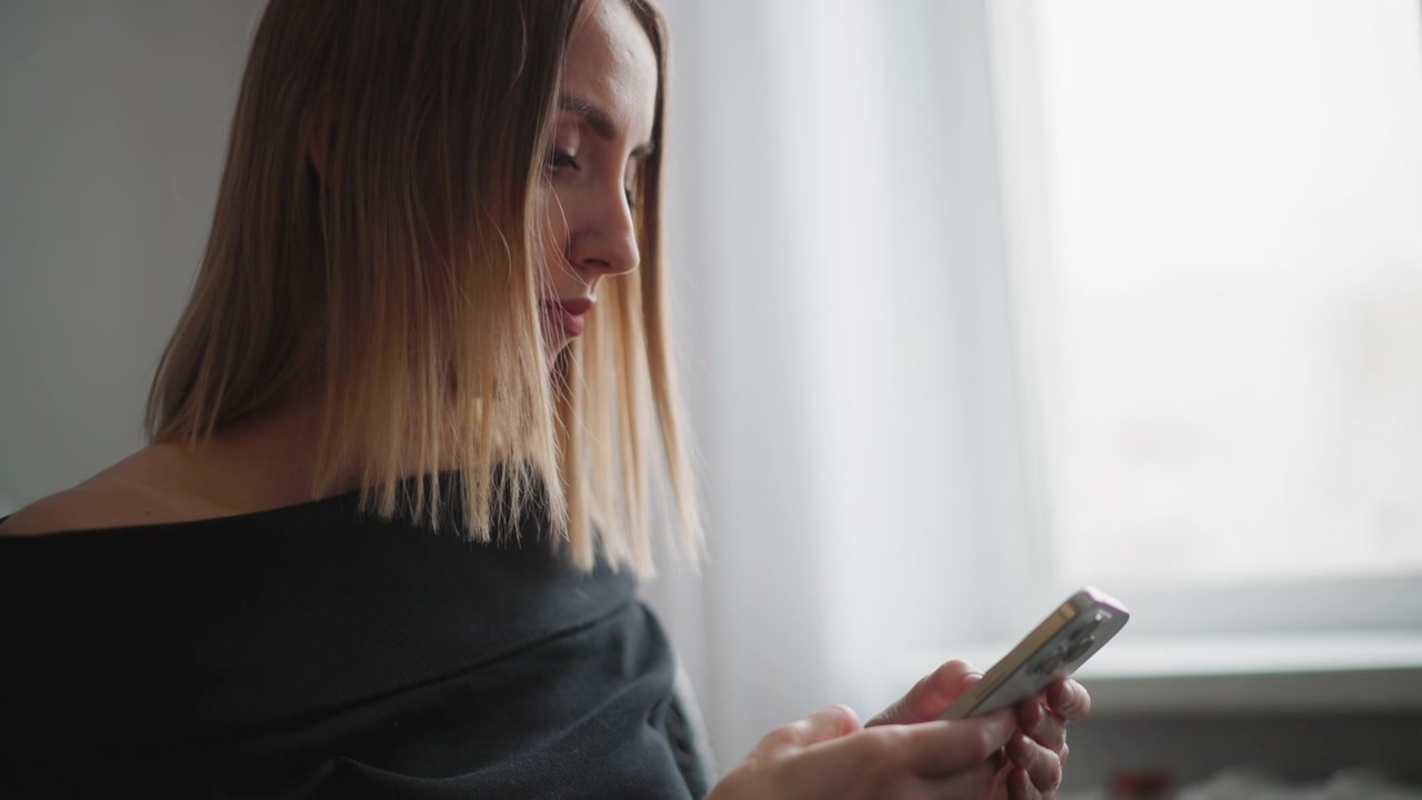 Primo piano di una donna con capelli lisci che guarda il suo smartphone in una luce naturale soffusa. La donna indossa un top nero a spalle scoperte, concentrandosi sul dispositivo mentre è seduta vicino a una finestra con luce diurna intensa