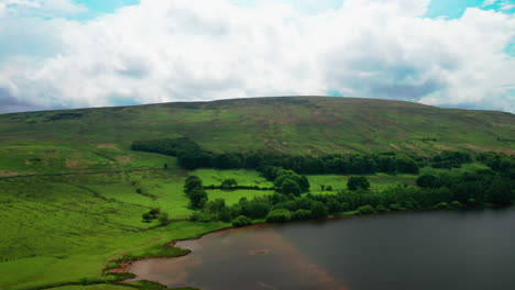 Aerial-dropping-shot-showing-a-still-lake-situated-at-the-bottom-of-a-hill,-bright-sunny-day