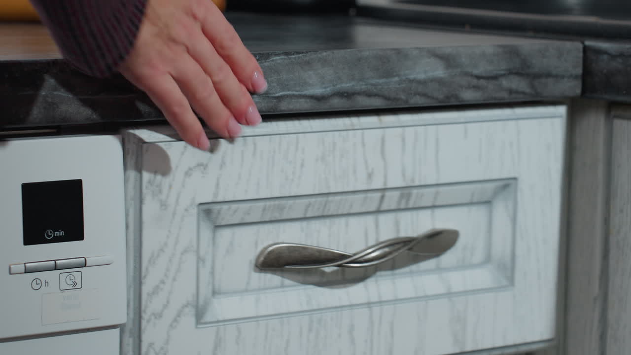 Close-up of hand in plaid clothing opening kitchen cabinet drawer, returning spoon with neatly polished nails, modern kitchen with white cabinetry, dark countertop, built-in dishwasher