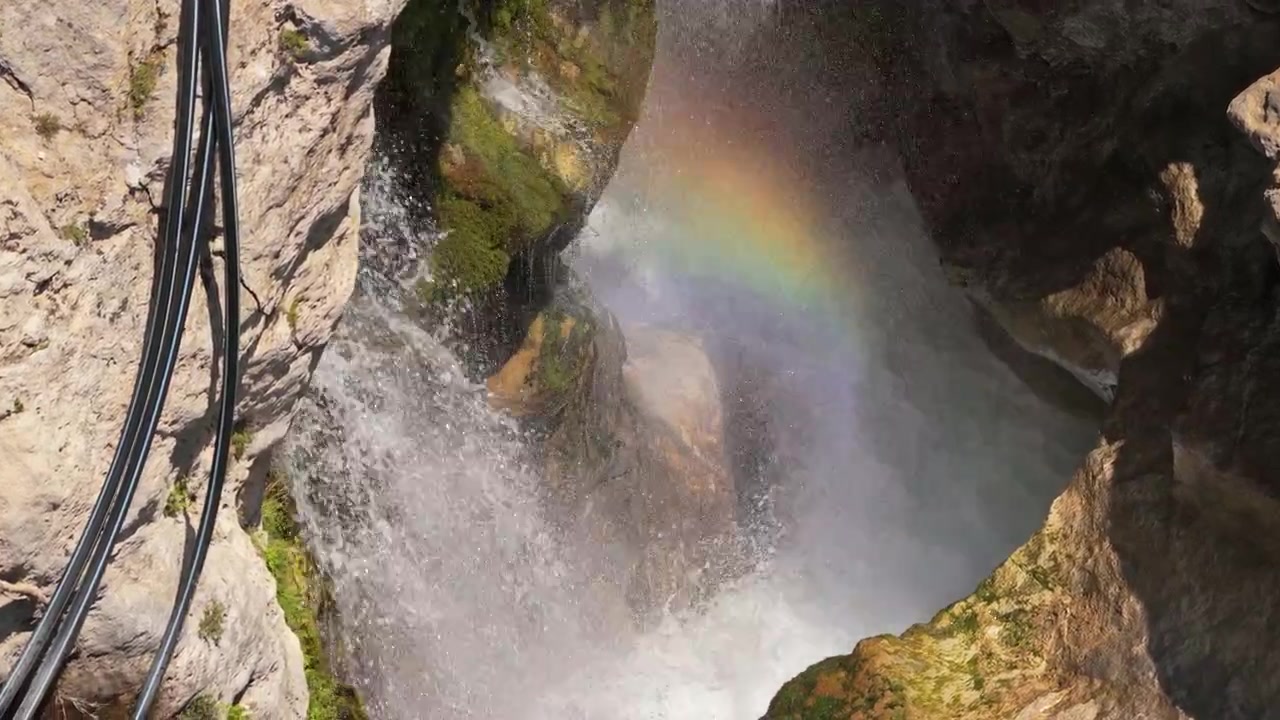 Luftfoto med drone av en foss som stuper ned i en tåkete steinete kløft med en regnbue i spruten, Kourtaliotiko-kløften, Hellas