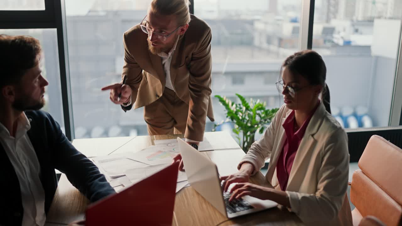 A blond man in glasses with a beard tells what his colleagues need to do during a meeting at work in a modern office. Office workers discussing new ideas in a modern office with a panoramic window
