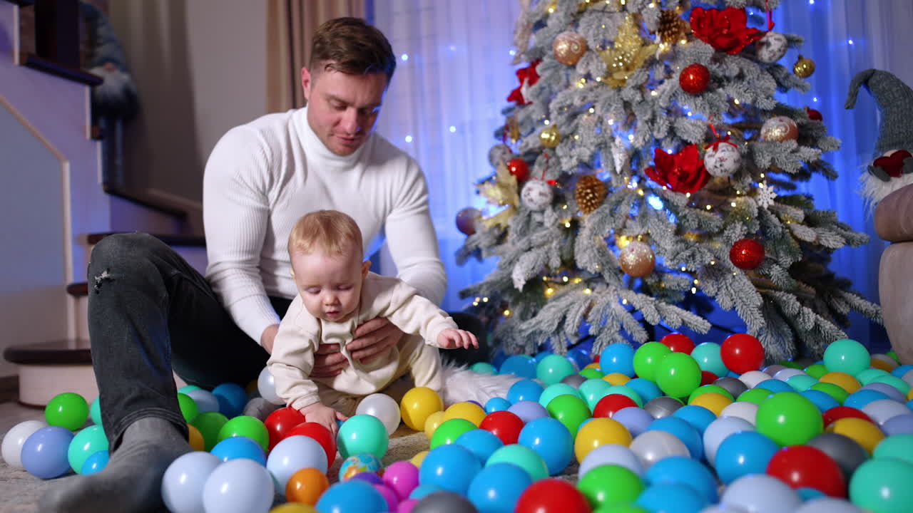 Father and his little infant son sit on the floor near the Christmas tree. Dad helps his baby to stand up among the multiple plastic balls. Black cat jumps at foreground.