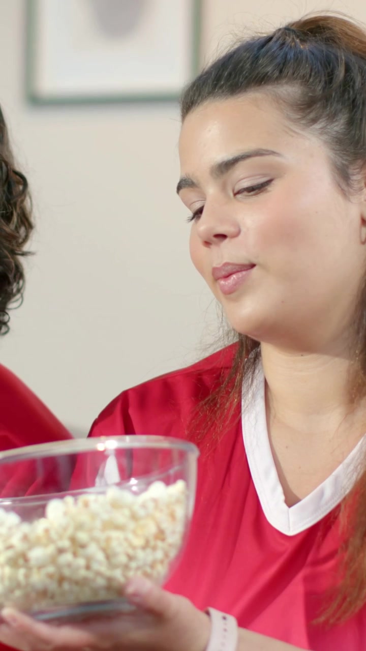 Video verticale: Mano che raggiunge una donna in maglia rossa che sposta una ciotola sul divano, offrendo popcorn.