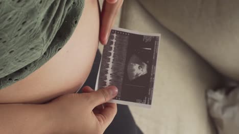 mujer embarazada se frota el vientre mientras sostiene una imagen de ultrasonido, vista desde arriba