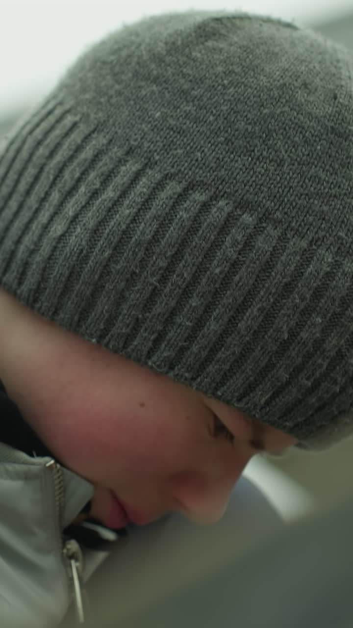 Close-up of a young boy wearing a gray jacket and green beanie, deeply focused on a task near an iron railing, he looks frustrated