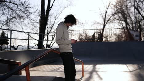 Side-View-Of-A-Young-Curly-Headed-Male-Skateboarder-Browsing-In-His-Mobile-Phone-In-The-Skate-Park-On-A-Sunny-Day