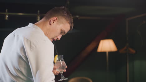 billiard player in white shirt sipping lemon drink with black straw in dimly lit billiard room. overhead lights cast dramatic shadows on green pool table. focused expression, relaxed posture setting