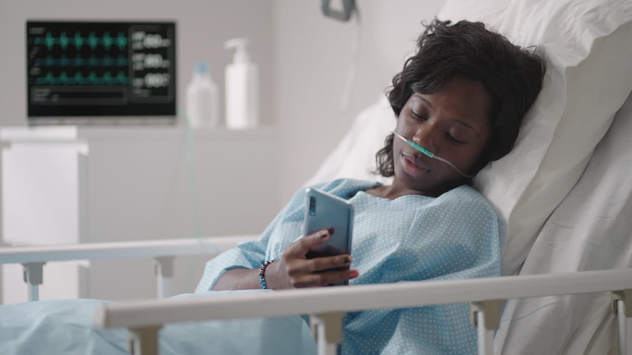 Black woman Patient in Hospital with Saline Solution Volumetric Infusion Pump using mobile phone on examination couch. African women lying in hospital bed with smart mobile phone while in hospital.