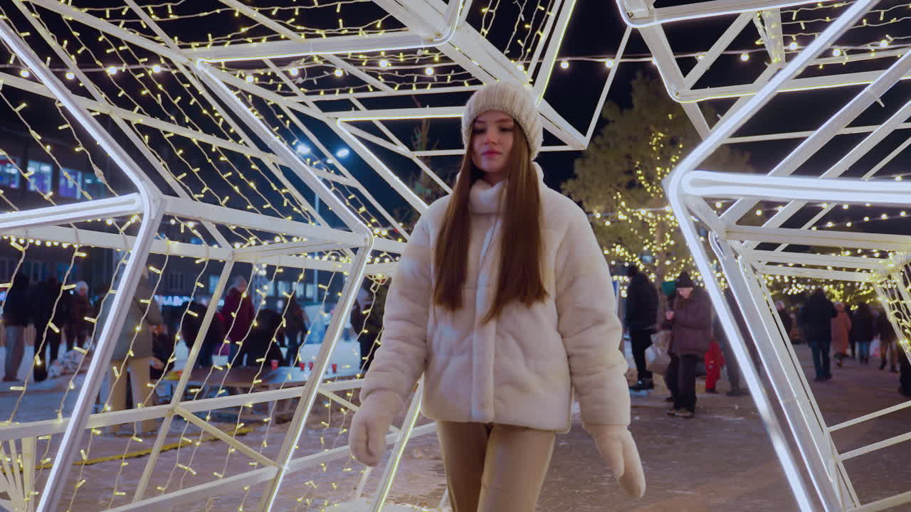 Young woman in cozy winter outfit walks through large illuminated star decoration at night, festive lights create a magical atmosphere as people gather in the background