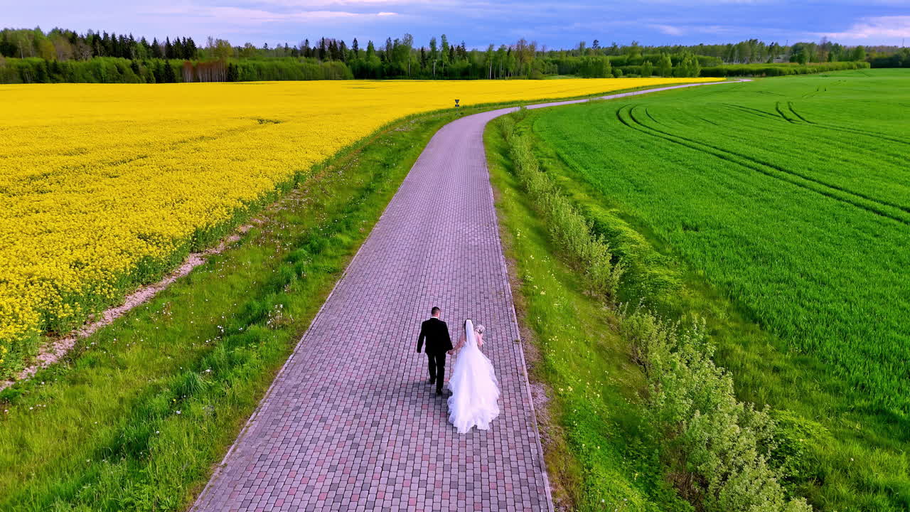 A couple in wedding attire walks hand in hand down a rural brick road lined with yellow blooms and green fields, passing a parked black car under a wide spring sky