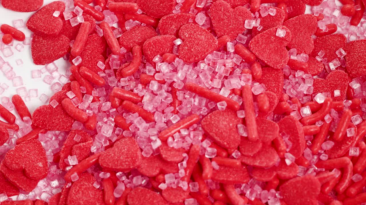 Red heart-shaped candies and sprinkles cascade onto a white surface, creating a vibrant and playful visual effect