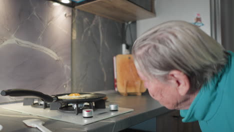 close-up shot of older woman observing eggs cooking in frying pan on stove, focused on food preparation in modern kitchen with stovetop, utensils, and kitchen tools visible