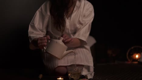 beautiful brunette in bathrobe taking teapot and pouring tea in cup drinking herbal tea at the spa in slow motion. thai tea