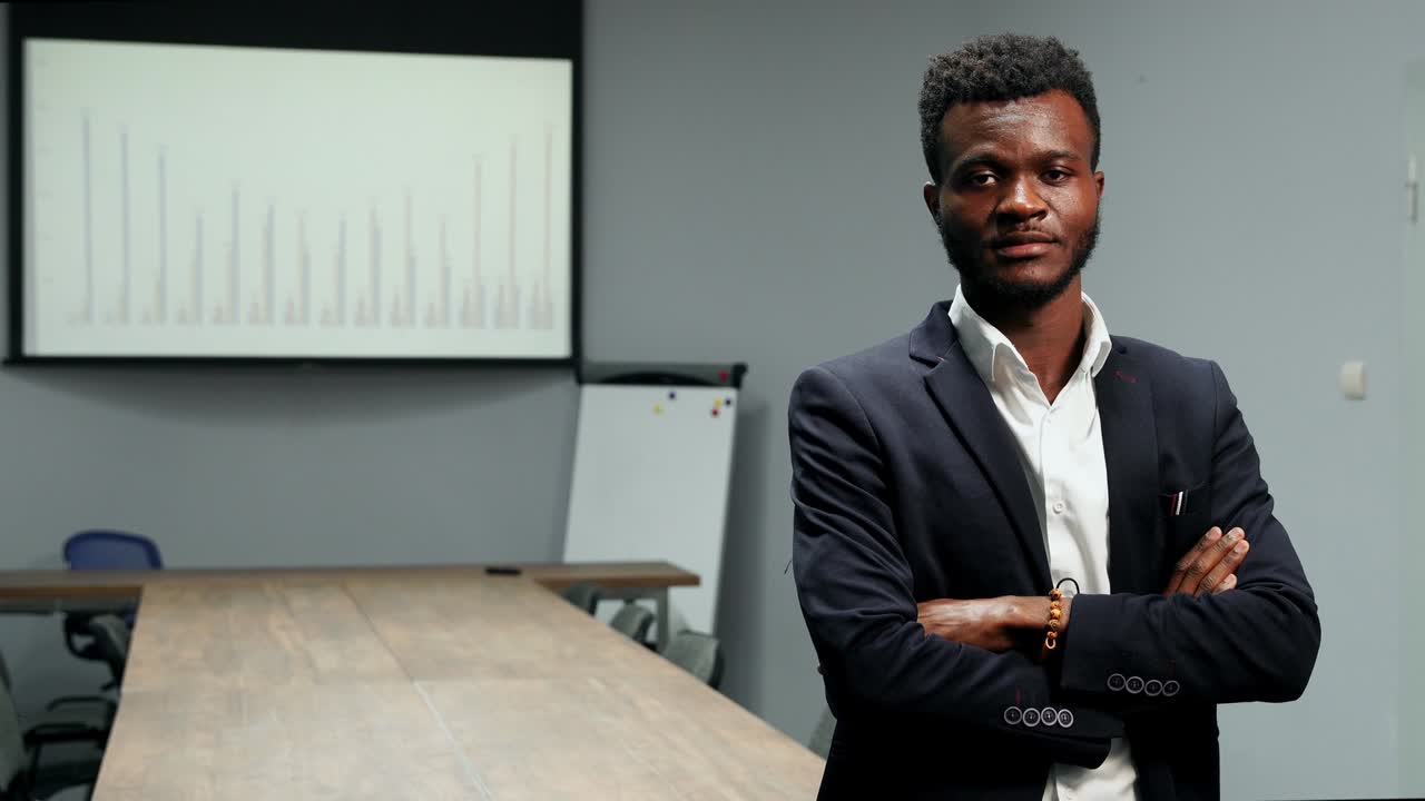 retrato de un hombre de negocios afroamericano en traje y camisa blanca de pie en la oficina en el fondo de una gran mesa y gráficos en la pantalla mirando a la cámara