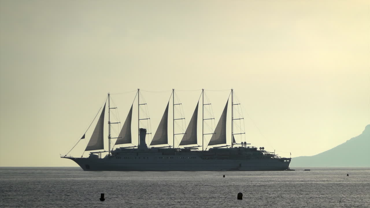 Distant view of a large multi-mast sailing ship gliding across the Mediterranean Sea at sunset with mountains on the background