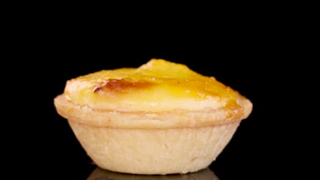A single egg tart dessert rotates smoothly on a glossy black surface, illuminated by soft studio lighting, highlighting its golden baked texture and pastry details