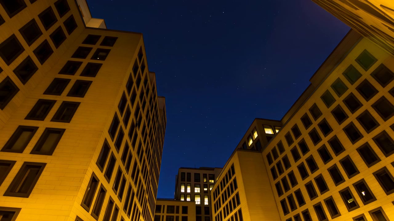 Vista dal basso verso l'alto di complessi di edifici per uffici a media altezza che racchiudono un cortile centrale, con luce diurna mutevole, finestre luminose, riflessi del vetro e forti linee verticali contro il cielo aperto.