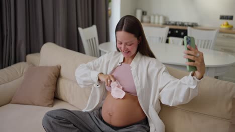 A-happy-pregnant-girl-is-vlogging-using-her-smartphone-about-her-pregnancy-and-expecting-a-baby-showing-little-socks-near-her-belly-on-the-sofa