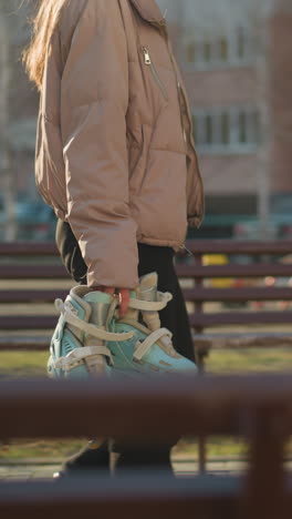 una persona vestida con una chaqueta de melocotón y pantalones negros camina a través de un parque iluminado por el sol, sosteniendo un par de patines en una mano. bancos y edificios residenciales son visibles en el fondo