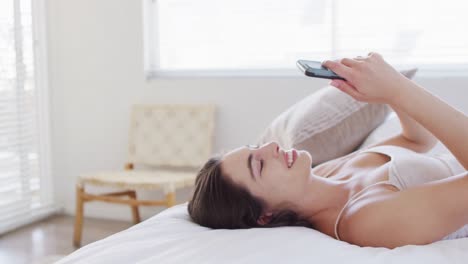 happy caucasian woman relaxing, lying in bed and using smartphone