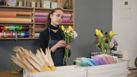 Young-female-florist-arranging-modern-bouquet-at-flowers-shot-standing-by-the-table.-She-combines-creamy-roses-and-yellow-tulips