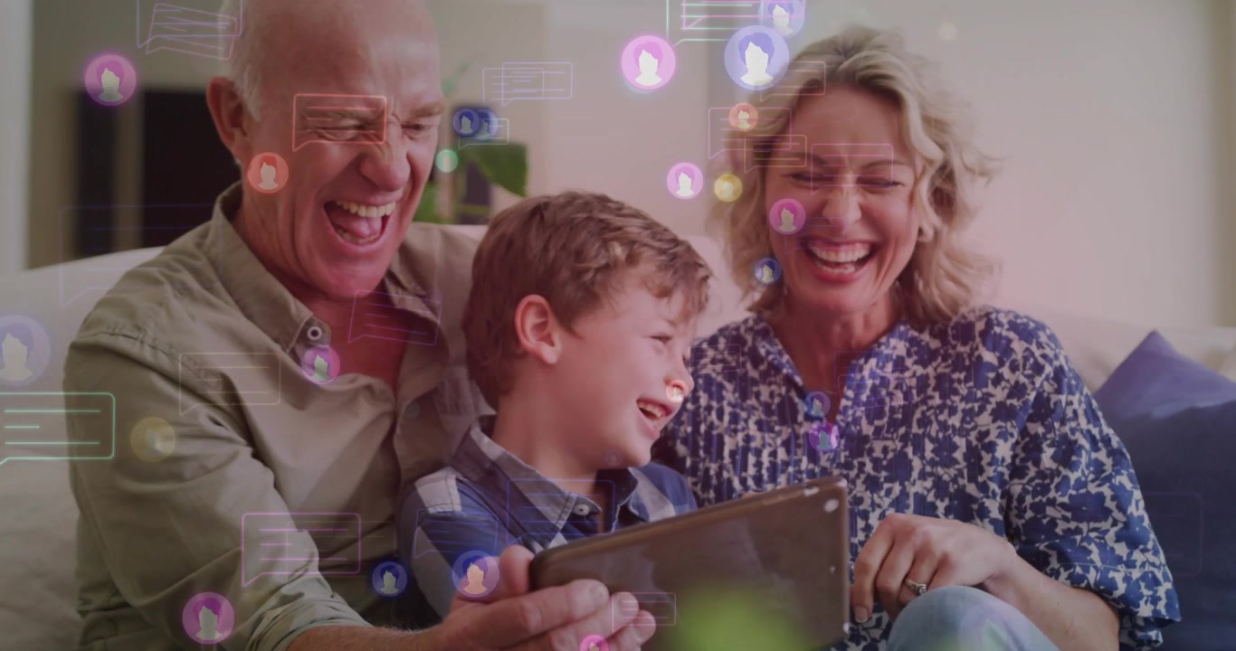 Nonni con il nipote seduti sul divano, il nonno che tiene un tablet, il bambino che reagisce alle icone tecnologiche.