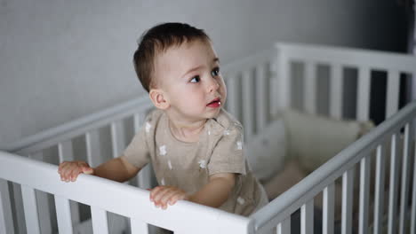 active little boy stands in his crib. kid holds by the handrails and jumps excitedly shaking the bed.