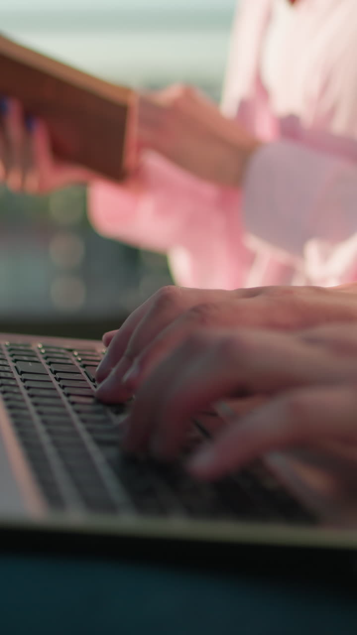 primer plano de manos escribiendo en un teclado de portátil al aire libre mientras otra persona sostiene un libro en el fondo, el fondo borroso presenta vegetación y luz solar