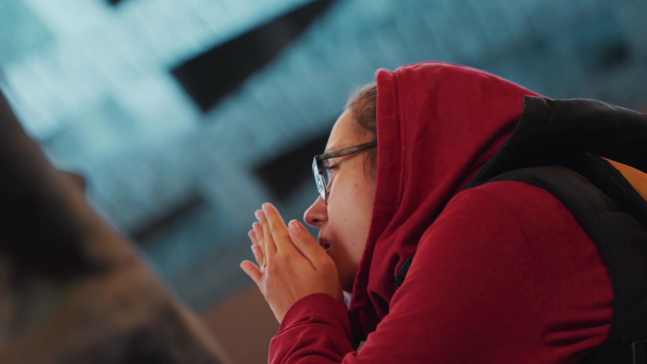 Night street closeup woman gazing upward contemplative pause student traveler hands clasped eyeglasses red hooded jacket cool blue bokeh lights urban architecture introspective mood