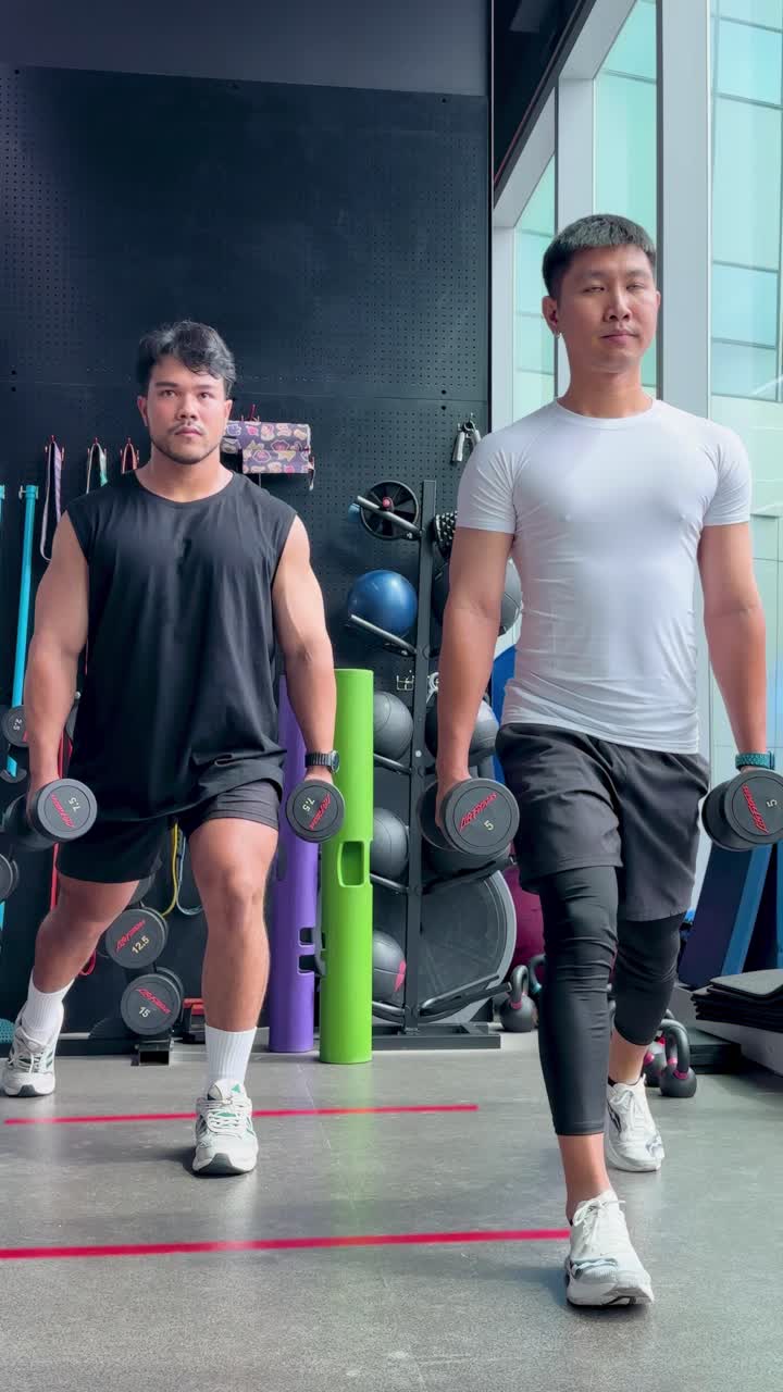 Dua pria melakukan dumbbell lunge secara sinkron di lingkungan gym yang terang benderang, menunjukkan kekuatan dan koordinasi.