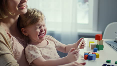 Madre-Joven-Sosteniendo-A-Una-Niña-Pequeña-Llorando-En-Su-Regazo-Y-Mostrando-Algo-En-El-Teléfono-Móvil-Mientras-Intentaba-Calmarla