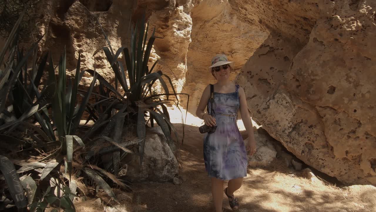 A backwards dolly camera movement of a young girl wearing a sun hat, dress, sunglasses and a camera, on an adventure exploring the rocky landscape on a sunny summer day in Cyprus.