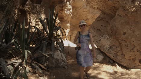 a backwards dolly camera movement of a young girl wearing a sun hat, dress, sunglasses and a camera, on an adventure exploring the rocky landscape on a sunny summer day in cyprus.