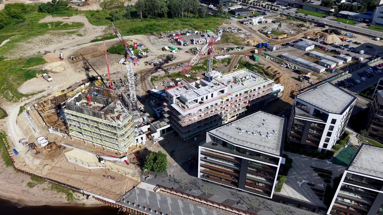 Dynamic aerial footage shows cranes and workers at a riverside construction site, with new modern buildings rising alongside water, vibrant sunlight, and bustling urban landscape. Aerial footage.
