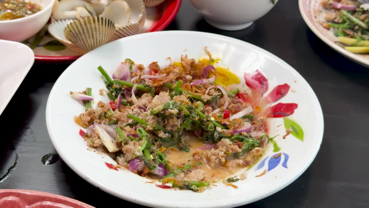 A hand uses a spoon to serve spicy minced pork salad from a decorated plate, under bright lighting on a restaurant table with side dishes